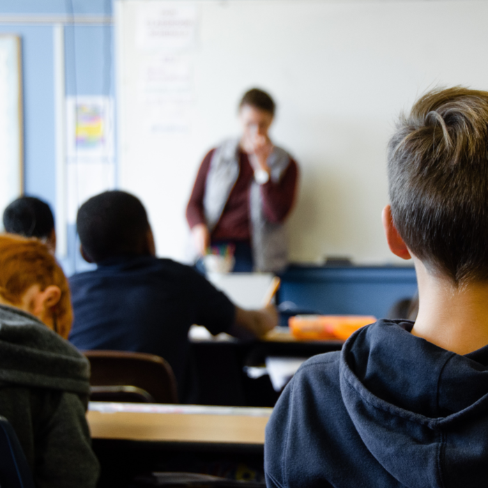 Schüler*innen sitzen an Tischen und schauen zur Lehrperson.