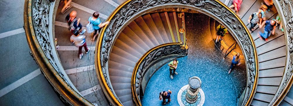 A large spiral-staircase decorated with stucco, photographed from above. People are walking up and down the stairs.