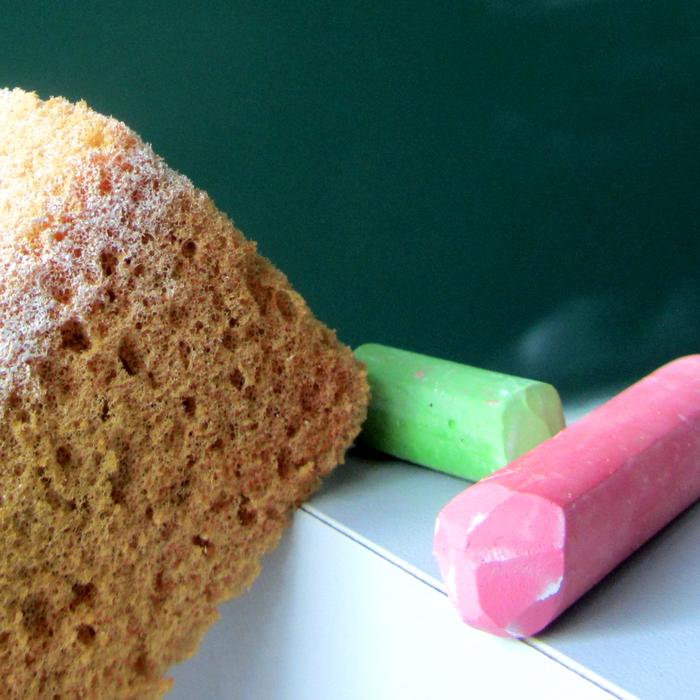 A sponge, a green stick of chalk and a pink stick of chalk in front of a blackboard.