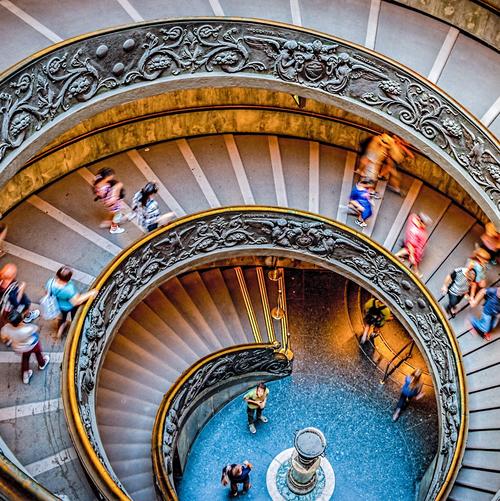 A large spiral-staircase decorated with stucco, photographed from above. People are walking up and down the stairs.
