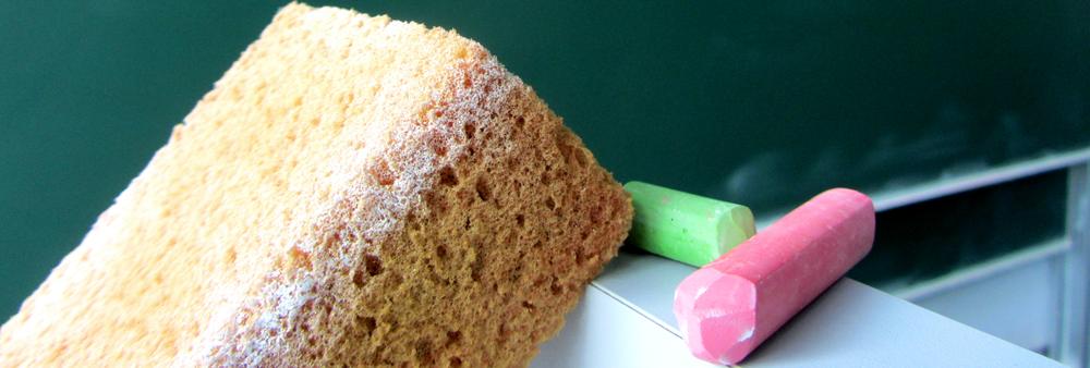 A sponge, a green stick of chalk and a pink stick of chalk in front of a blackboard.